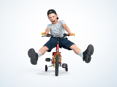 A happy, screaming boy in a T-shirt, cap and shorts rides a bicycle directly to the camera with his legs raised, isolated on a light blue background. File contains a path to isolation.
