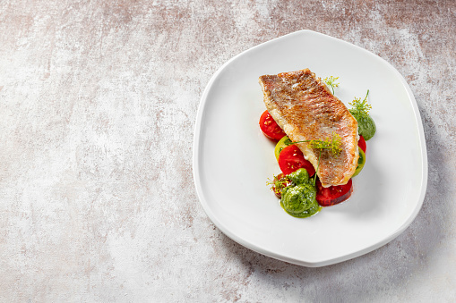 Grilled sea bass fillet with green and red tomatoes and pesto sauce. Food lies on a light ceramic plate on a light background.