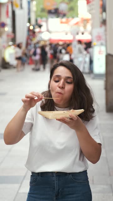 Hispanic young woman eating takoyaki at Dotonbori street in Osaka, Japan.