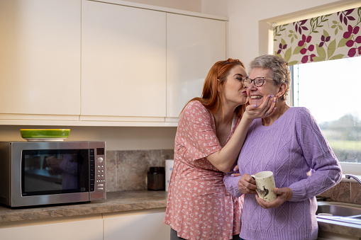 Waist-up shot of a granddaughter who is pregnant and grandmother standing together in a kitchen. The granddaughter is kissing and holding her grandma on the cheek. They are both wearing casual clothing.