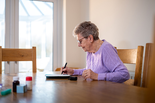 Side view shot of a senior female adult checking her pulse trace using a pulse oximeter. She is wearing a purple jumper, sitting at her dining table, looking down at the pulse oximeter reading. She has her medication on the dining table in front of her.