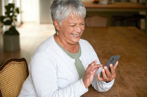 Senior woman sitting by the dinner table in the kitchen holding her phone, laughing