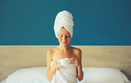 Happy young woman enjoying with cup of coffee drying her wet hair after bath with wrapped towel on her head sitting on bed in the morning