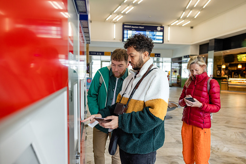 A medium close side view of a same sex couple who are travelling Europe and are visiting Garmisch in Germany. They are in the towns train station and are paying for their onward travel using the contactless and cashless ticket machines.