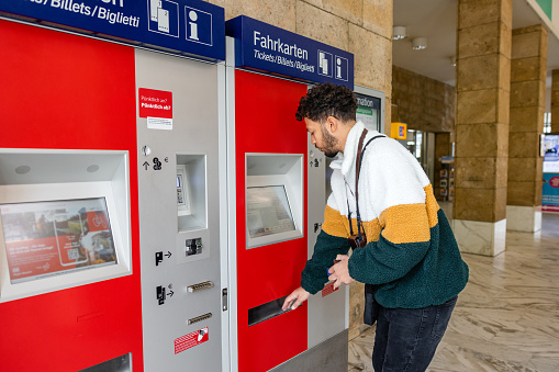 A medium close up of a man who is from the UK and travelling in the European Alps in Garmisch train station in Germany. He is navigating his way around the ticket machine and paying for and collecting them from the machine.