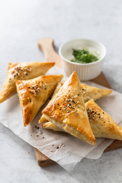 Filo pastry triangle hand pies on baking paper Cheese stuffed triangle filo pastry savory pies served with yoghurt sauce over baking paper on wooden board spanakopita stock pictures, royalty-free photos & images