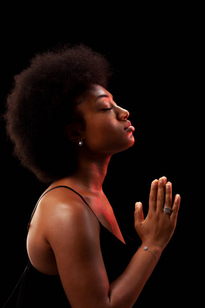 Studio portrait of elegant african american lady with curly hair afro hairstyle against black background. Girl in black dress praying. stock photo