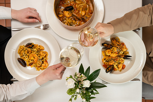 Homemade pasta with shrimp, salmon, mussels in a wine-cream sauce in a copper dish with a handle and baked in the oven under a dough lid. Place the dishes on a table with a light tablecloth. The waiter uses a fork and a spoon to place pasta and white plates.