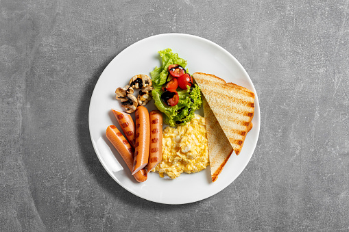 Scrambled eggs with sausages and toast, lettuce, cherry tomatoes and mushrooms in balsamic sauce. Food lies on a light ceramic plate on a gray stone background.