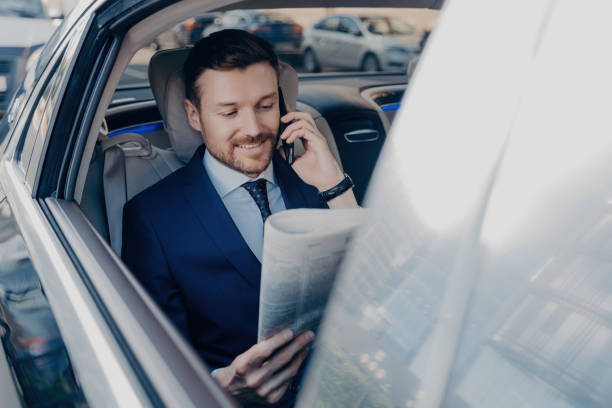 Happy executive manager in stylish formal wear multitasks: reads newspaper, speaks on smartphone with partner while stuck in traffic.