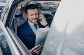 Happy executive manager in stylish formal wear multitasks: reads newspaper, speaks on smartphone with partner while stuck in traffic.