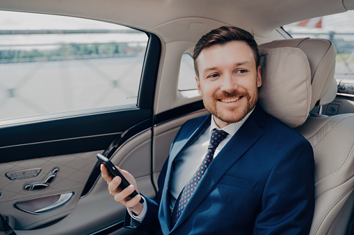 Cheerful executive in elegant tuxedo checks stock prices on cellphone, rides to financial meeting in luxury car with driver.