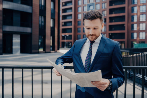 Focused businessperson in suit checks newspaper for interesting news, stands alone outdoors, leaning on black fence by office plaza buildings, enjoying work break. Focused businessperson in suit checks newspaper for interesting news, stands alone outdoors, leaning on black fence by office plaza buildings, enjoying work break. business person one man only blue standing stock pictures, royalty-free photos & images