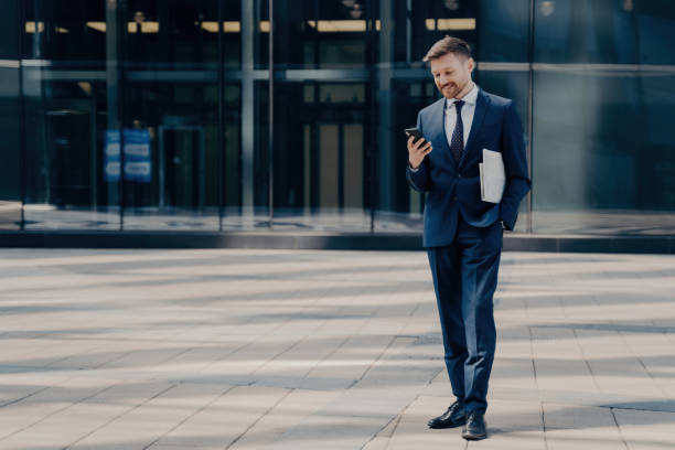Cheerful employee in blue suit texts/chat on smartphone, holds newspaper, stands next to glass building outdoors. Business people and technology concept. Cheerful employee in blue suit texts/chat on smartphone, holds newspaper, stands next to glass building outdoors. Business people and technology concept. tie game stock pictures, royalty-free photos & images