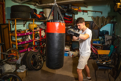 Caucasian teenage boy kicking the punching bag at the messy garage