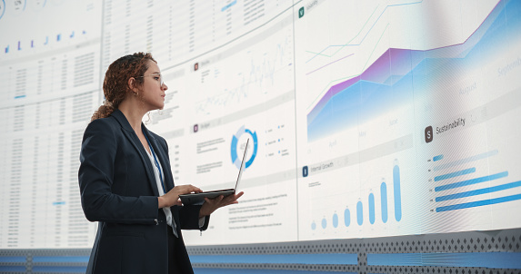 In the Monitoring Office: Successful Hispanic Businesswoman Holding Laptop Computer And Analyzing Data On Big Digital Screen With Graphs and Charts. Female IT Entrepreneur Running Software Company.