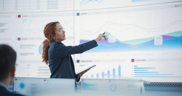 Hispanic Female Senior Data Scientist Reviewing Reports Of Risk Management Department On Big Digital Screen In Monitoring Room. Diverse Consulting Company Employees Working Behind Desktop Computers. científica de datos senior hispana revisando informes del departamento de gestión de riesgos en una pantalla digital grande en la sala de monitoreo. diversos empleados de empresas de consultoría que trabajan detrás de computadoras de escritorio.