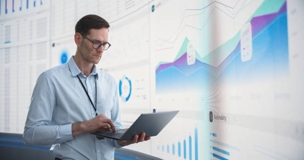 istock Caucasian Male Business Analyst With Laptop Computer Standing Next To Big Digital Screen With Graphs And Charts. Successful Man Assessing Risks And Developing Strategies For Company Growth, Expansion. 1522425224