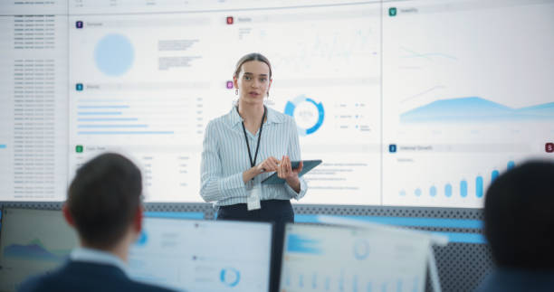istock Female Project Manager With Tablet Computer Is Giving Presentation To Diverse Employees In Front of Big Digital Screen In Monitoring Room. Team Of Data Scientists Listening To New Objectives, Tasks. 1522425343