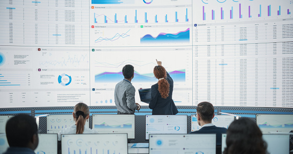 Wide Zoom In Back Shot Of Diverse Male And Female Risk Managers Analyzing Data On Digital Screen In Monitoring Room. Multiethnic Employees Working On Computers For Succesful Venture Capital Company