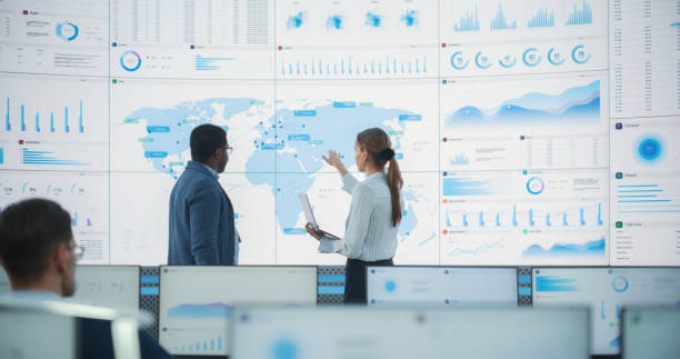 istock Back View Of Black Male Data Scientist Talking To Caucasian Female Supervisor In Front Of Digital Screen With World Map In Monitoring Office. Employees Working Behind Computers in Distribution Company 1522425663