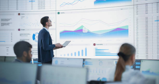 istock Male Senior Data Analyst Reviewing Financial Information And Reports on Big Digital Screen in Monitoring Office. Diverse Employees Working Behind Desktop Computers In Venture Capital Company. 1522425831
