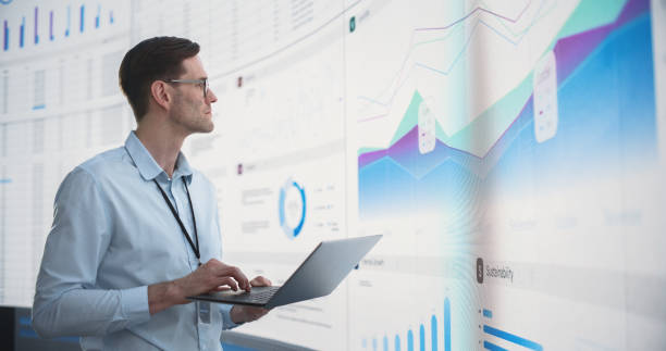 istock Caucasian Male Business Analyst Holding Laptop Computer And Standing Next To Big Digital Screen With Graphs And Charts In Office. Successful Man Reviewing Financial Reports For Venture Capital Company 1522425986