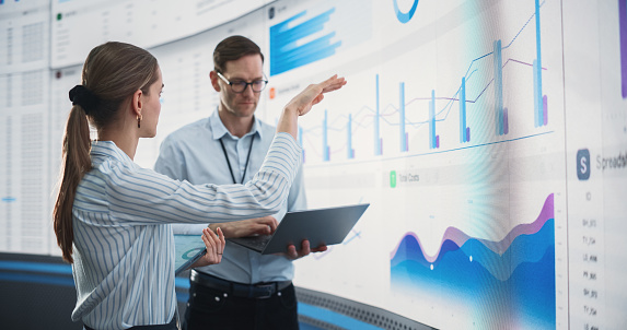 Caucasian Male And Female Data Scientists With Laptop And Tablet Standing Next To Big Digital Screen With Graphs And Charts In Monitoring Office. Colleagues Discussing Business Opportunities, Risks.