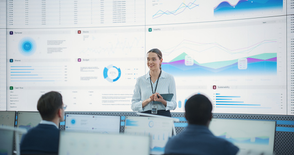 Caucasian Female Project Manager With Tablet Computer Is Giving Presentation To Diverse Employees In Front of Big Digital Screen In Monitoring Room. Team Of Data Scientists Listening To New Objectives