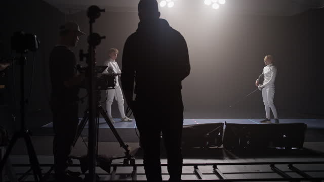 Fencing match recorded in a professional studio. Camera rolling. Two fencers on a stage filmed by the crew