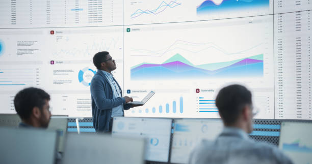 istock Male Financial Data Analyst Using Laptop And Studying Information And Graphs On Big Digital Screen In Monitoring Office. Black Man Working For Big Consulting Firm, Diverse Employees Behind Computers. 1522430292
