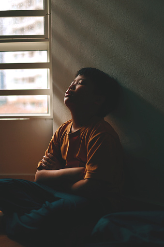 In the shadows of a home corner, a little Asian boy with a sad and lonely expression hides. His face is illuminated by the shadowy light coming through a window.