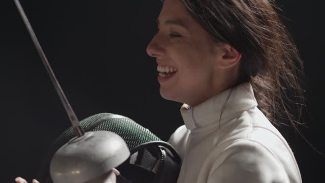 Close-up female fencer removing protective headgear then cheering and jumping with foil sword in hand. Celebrating victory