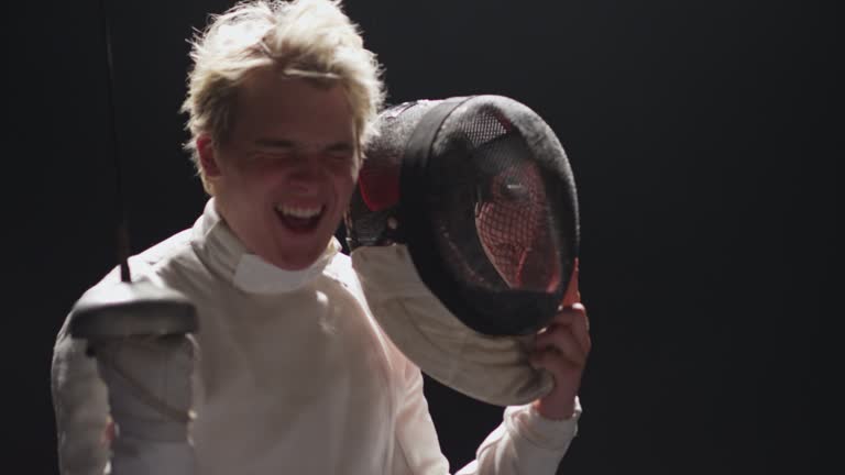 Male fencer removing protective headgear then cheering and jumping with foil sword in hand. Celebrating victory