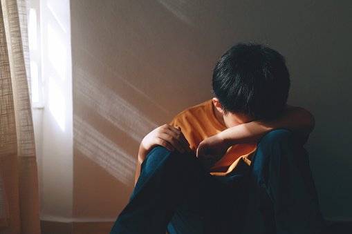 In the shadows of a corner in his home, a little Asian boy sits, looking sad and crying, with his head resting on his knees. The only part of him illuminated is his face, as the shadowy light filters through a window.