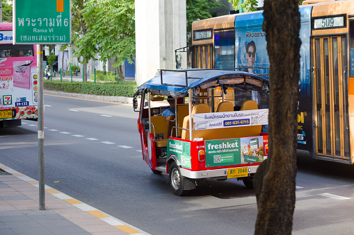 Electric modern thai tuktuk taxi driving on Phahonyothin Road in Bangkok Phaya Thai. A bus is overtaking tuktuk