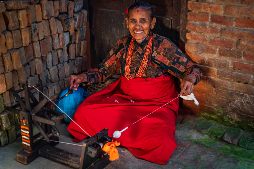 Old Nepali lady sitting near the Bhaktapur Durbar Square and spinning the wool. Bhaktapur is an ancient town in the Kathmandu Valley and is listed as a World Heritage Site by UNESCO for its rich culture, temples, and wood, metal and stone artwork.