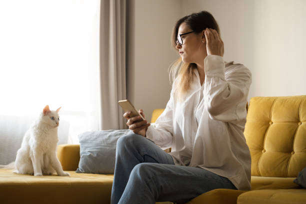 istock Attractive middle-aged brunette woman wearing eyeglasses using mobile phone looking away with beautiful white cat,sitting on yellow sofa at home.Freelance, working online with pet.Copy space for text 1522869545