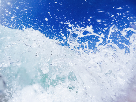 A closeup view of wave splashes. The wave depicted is light blue and foamy, generating numerous splashes that are suspended in the air. The background showcases a vibrant blue sky without any clouds, providing a contrasting backdrop to the dynamic scene.