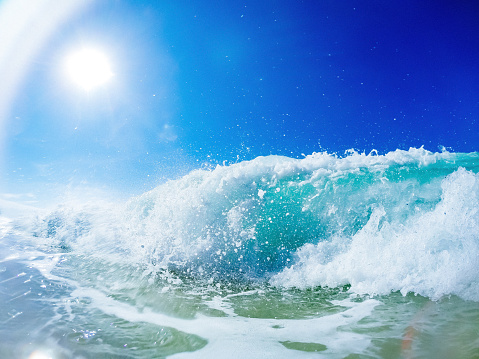 Close up photograph of a wave crashing onto the beach. The wave, adorned in a foamy texture and light blue hue, rises to its peak. Taken from a ground perspective, the image reveals numerous splashes suspended in the air. The vibrant blue sky, unobstructed by clouds, is illuminated by the radiant sun, casting a direct light towards the camera.