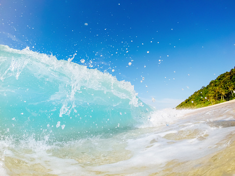Close up photograph captures the moment a foamy light blue wave crashes onto the beach. The shot, taken from a ground perspective, showcases the wave at its peak, creating splashes in the air. The background features a clear, vibrant blue sky without any clouds.