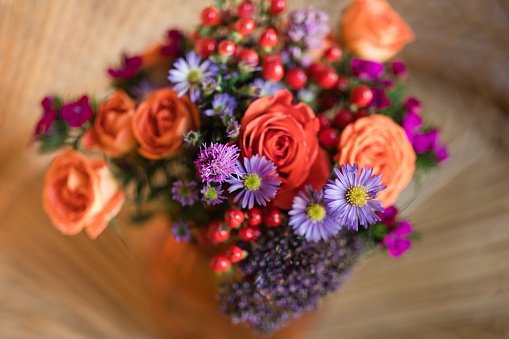 A Colorful Summer Floral Arrangement of Coral Roses, Purple Daisies, Red Berries & Other Flowers in an Orange Mason Jar Glass Vase Sitting on a Boho Vintage Rattan Peacock Chair in South Florida in the Summer of 2023.