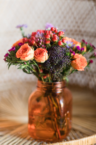 A Colorful Summer Floral Arrangement of Coral Roses, Purple Daisies, Red Berries & Other Flowers in an Orange Mason Jar Glass Vase Sitting on a Boho Vintage Rattan Peacock Chair in South Florida in the Summer of 2023.