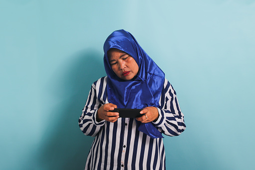 A bored middle-aged Asian woman in a blue hijab and a striped shirt is playing a game on her mobile phone, gesturing reluctantly, looking sad, and losing the game, isolated on a blue background.