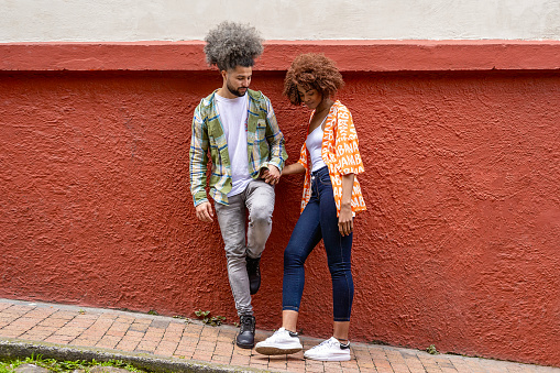 Happy latin american couple between 25 - 30 years old, talking leaning on a wall holding hands, the background is a red wall in the street of candelaria neighborhood in the city of bogota ( colombia ).