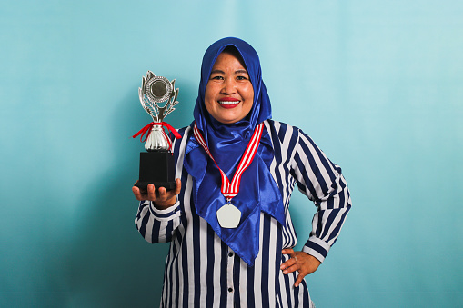 A happy middle-aged Asian businesswoman in a blue hijab and a striped shirt is showing an empty white medal while holding a silver trophy, celebrating her success, isolated on a blue background