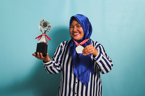 A happy middle-aged Asian businesswoman in a blue hijab and a striped shirt is showing an empty white medal while holding a silver trophy, celebrating her success, isolated on a blue background