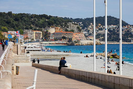 Nice, France - April 21, 2023: Mediterranean beach with the castle hill visible in the distance and buildings overlooking the sea. There are people on the beach enjoying the sunny weather