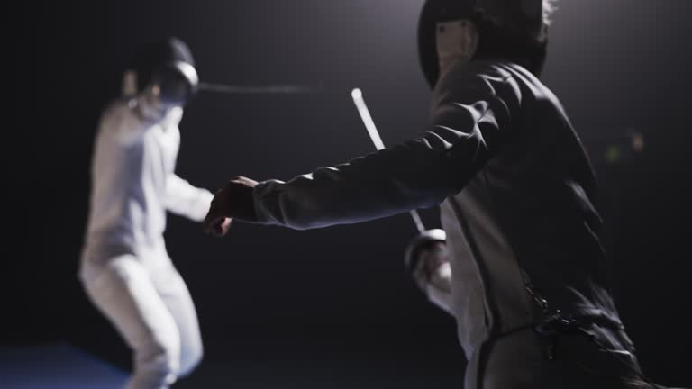 Rear view two professional fencers in full protective gear clashing foil sabers in a match. Attacking and ducking