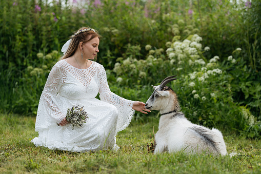 Young caucasian bride with dreadlocks petting white goat in countryside. Space for text.